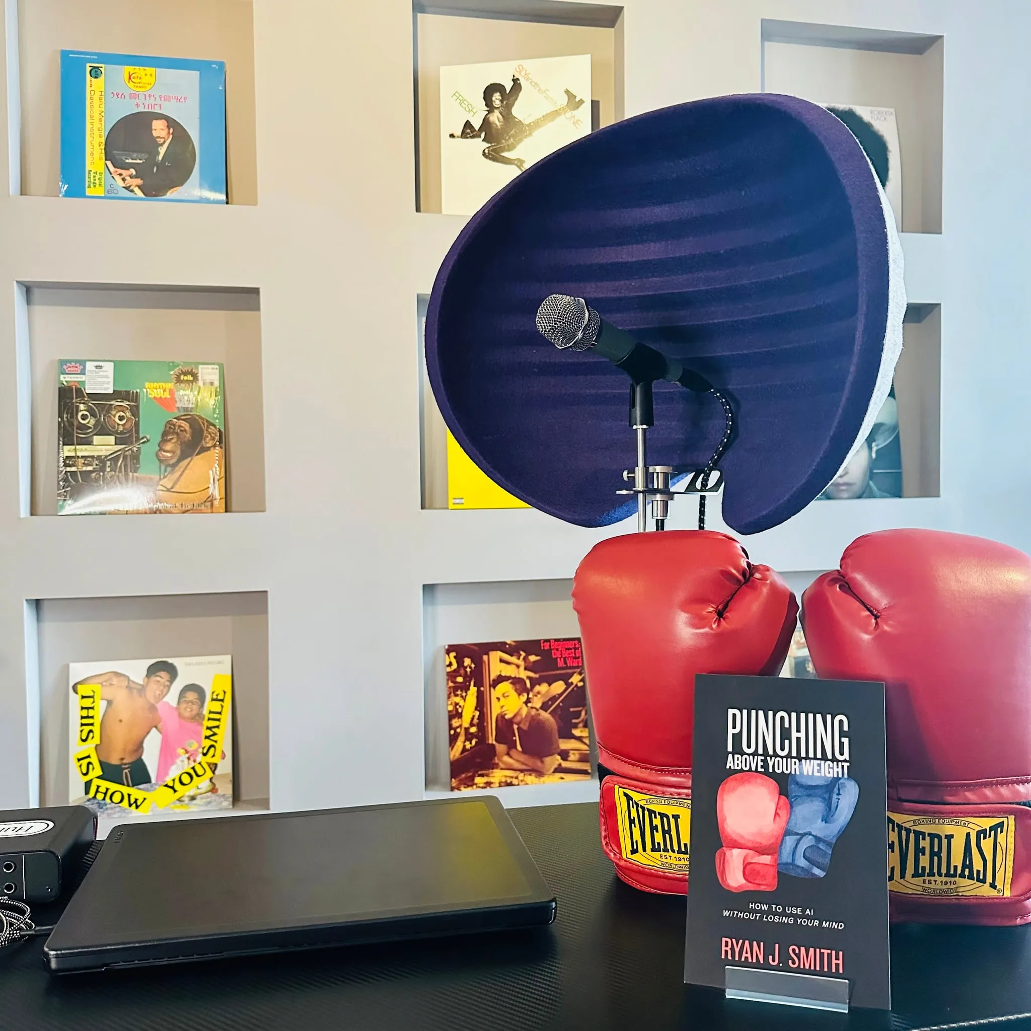 A microphone with a purple isolation shield on a desk, beside red boxing gloves and a copy of the book, with record shelves behind