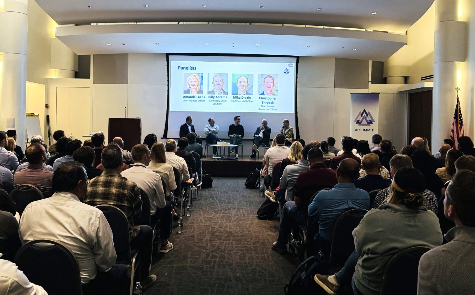 A full room at a corporate AI summit, watching a leadership panel